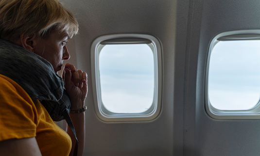 Mature, 55-years-old, Caucasian blonde woman, a tourist and traveler, flying in the airplane, sitting next to a window.