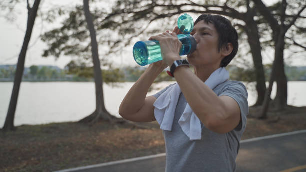 thirsty senior asian active woman drinking fresh water from bottle at trees park during evening sunset. mature female drink energy drinks refreshment hydration from tired and exhausted when workout. sediento mujer activa asiática mayor bebiendo agua dulce de botella en el parque de árboles durante la puesta del sol por la noche. madura hembra beber bebidas energéticas hidratación de refresco de cansado y agotado durante el entrenamiento.