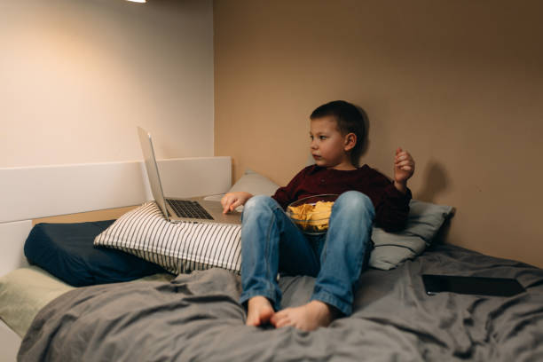 boy using laptop computer or digital tablet and eating junk food little boy eating unhealthy food. He is laying on bed and using laptop computer in his room terrible meal stock pictures, royalty-free photos & images