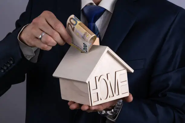 A businessman's hand lowers a 200 euro bill into a hole on the roof of a simolis wooden piggy bank house