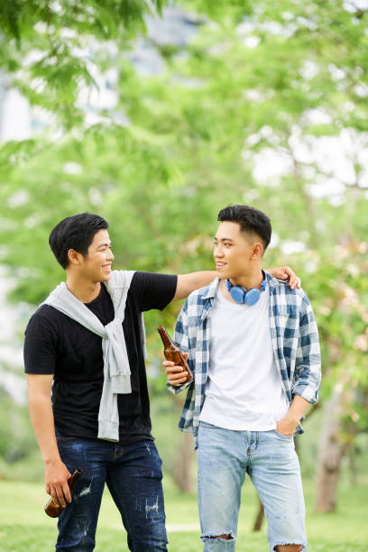 Two Asian young men drinking beer and talking to each other during their walk in the park