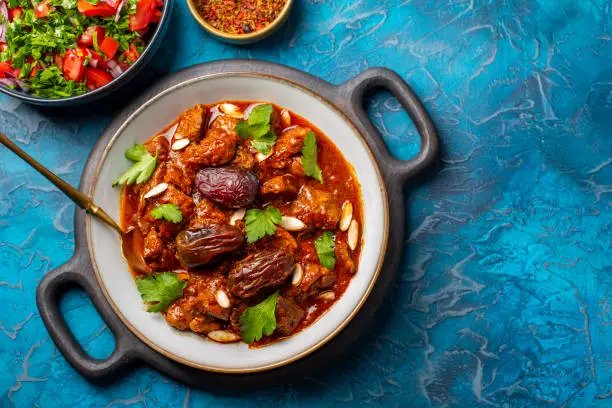 Traditional Moroccan lamb tagine simmered in spices, with dates and almonds. Salad and spices. Blue background.