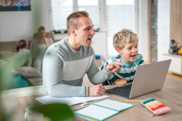Father and son during homeschooling Young adult man and a little boy using a laptop at the table. Mother and a child in the background sitting on a sofa. elementary student pointing stock pictures, royalty-free photos & images