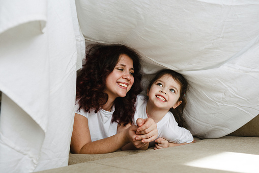 mom and little daughter play together on the couch hidden under a white blanket. joint games of mother and daughter