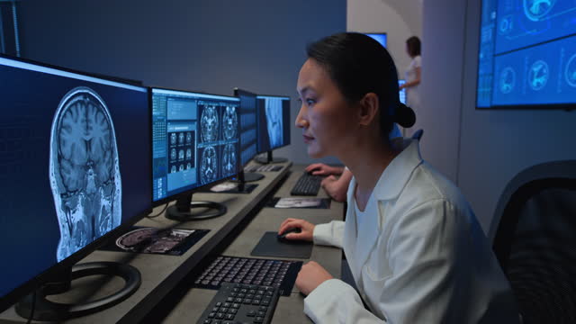彼女の男性の同僚と一緒に彼女のコンピュータ上のヘッドMRIスキャンをアナリーするアジアの女性放射線科医の広いドリーショット。スロベニアで撮影。