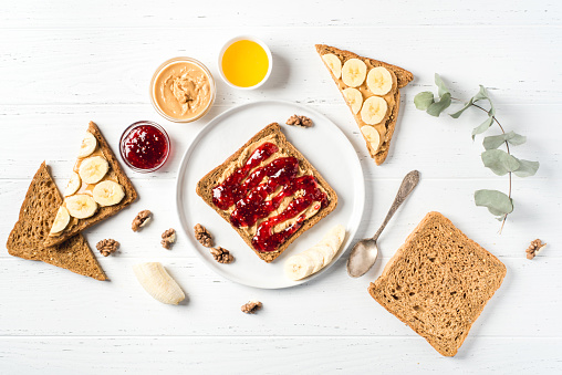 Homemade peanut butter sandwiches with raspberry jam and banana on white wooden background. Top view.