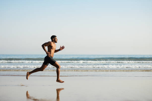 Powerful black man running barefoot by the sea Black fit man running barefoot by the sea on the beach. Powerful runner training outdoor on summer. barefoot stock pictures, royalty-free photos & images