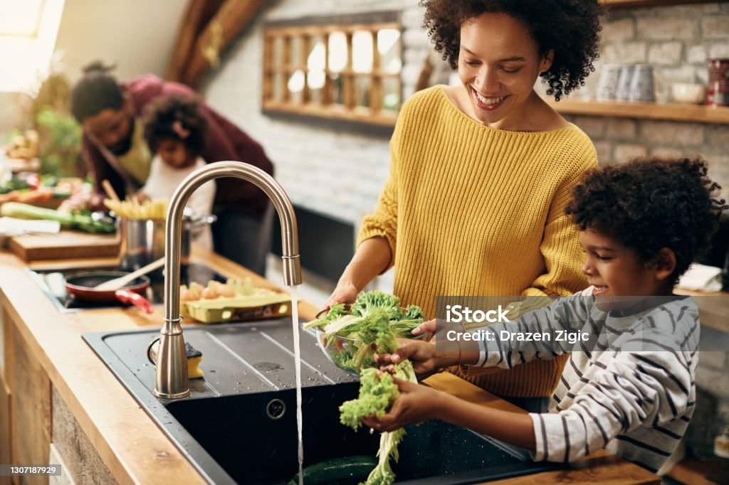 Happy black mother and son watching salad while preparing food in the kitchen. Happy African American boy assisting his mother and washing lettuce while preparing food in the kitchen. Focus is on mother. Family Stock Photo