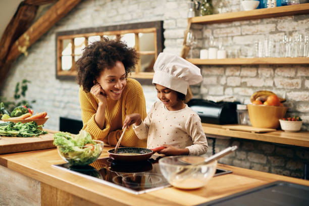 mère et descendant noirs heureux appréciant tout en préparant la nourriture dans la cuisine. - mother cooking daughter child photos et images de collection