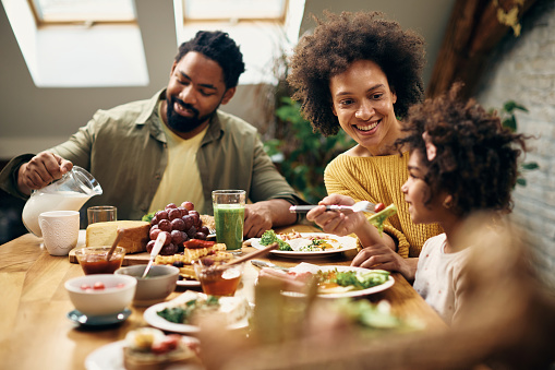 Happy African American mother feeding daughter during family meal at dining table.