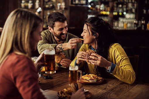 Happy man feeding his girlfriend with French fries while eating hamburgers in a pub. Focus is on woman.