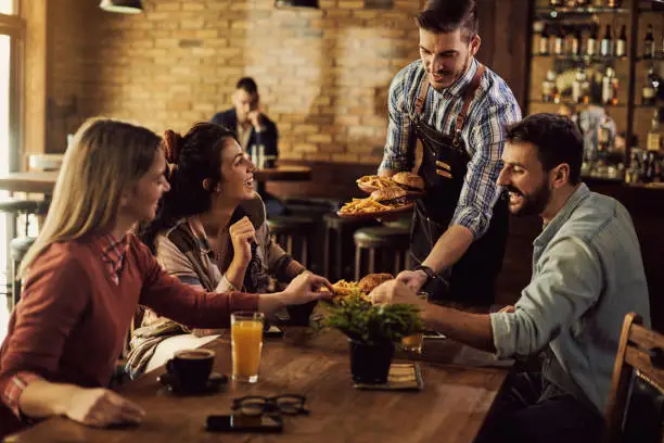 Photo of Happy waiter serving food to group of friends in a pub.