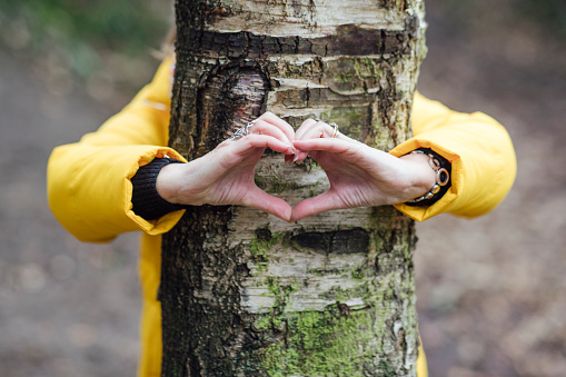 Senior woman with her arms around a tree making a heart with her hands. She is in the North East of England.