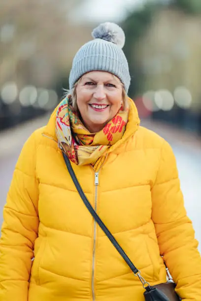 Portrait of a senior woman smiling while looking at the camera outdoors. She is wearing warm clothing in winter and is in the North East of England.