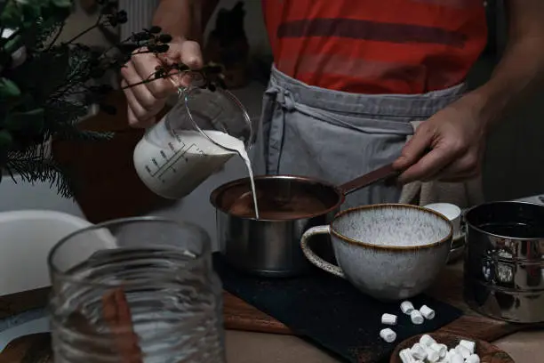A man in a gray apron pours milk into a saucepan. On a brown table with kitchen crockery. 
Frontal view.