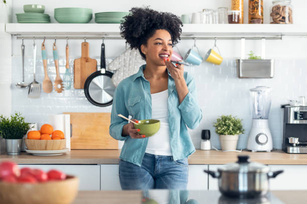 sorridente giovane donna afro che invia un audio con il suo cellulare mentre mangia noodles con bacchette in cucina a casa. - domestic staff audio foto e immagini stock