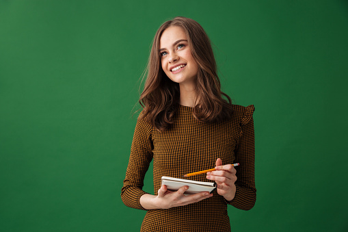 Image of cheerful young woman standing isolated over green background looking aside writing notes.