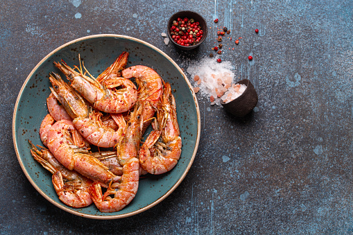 Whole fried big shrimps in blue bowl with salt and pepper on stone rustic background from above. Fresh cooked delicious grilled shrimps served on plate top view, healthy seafood meal. Space for text