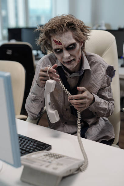 giovane uomo d'affari con il body painting zombie che tiene il ricevitore del telefono mentre è seduto alla scrivania in ufficio - frustration telephone computer it support foto e immagini stock