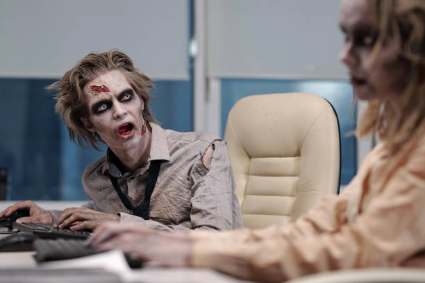 istock Zombie businessman with greasepaint on face and hands sitting by workplace and looking at co-worker 1307214566
