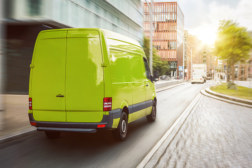 A green van in motion on an urban street. Buildings, a green tree and sunshine in the background.