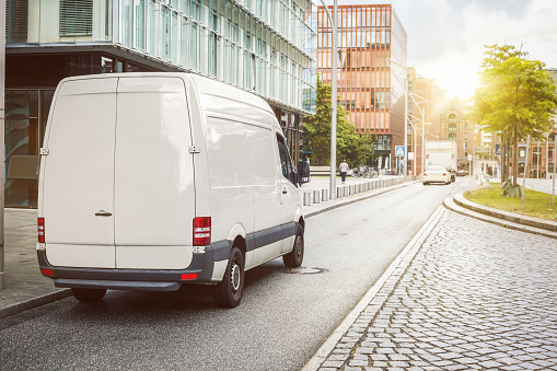 A white van in parked on an urban street. Buildings, a green tree and sunshine in the background.