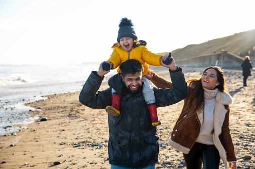 A multi-generation family wrapped up in warm clothing on the beach on a cold day in January. They are walking down the coastline and holding hands, the young boy is sitting on his father's shoulders.