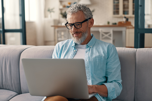 Senior man in casual clothing and eyeglasses using laptop and smiling while sitting on the sofa at home