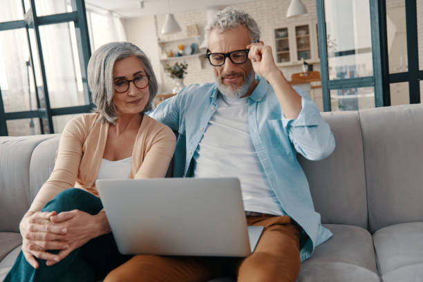 Modern senior couple in casual clothing Modern senior couple in casual clothing smiling and using laptop while bonding together at home mature couple stock pictures, royalty-free photos & images