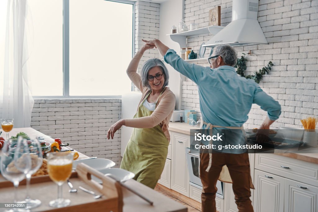 Beautiful playful senior couple Beautiful playful senior couple in aprons dancing and smiling while preparing healthy dinner at home Happiness Stock Photo