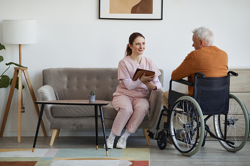 Full length portrait of smiling female nurse talking to senior man in wheelchair and using digital tablet at retirement home, copy space