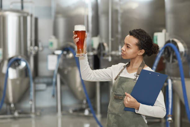 Young Woman Looking at Beer Glass at Brewery Waist up portrait of young African-American woman holding beer glass while inspecting quality of production at brewing factory, copy space microbrewery photos stock pictures, royalty-free photos & images