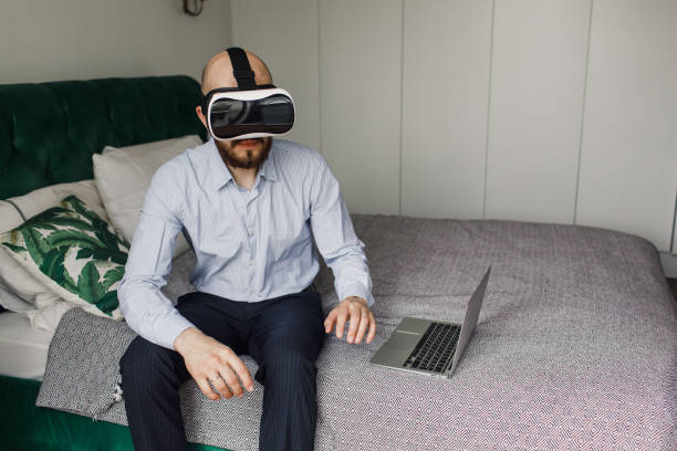 młody mężczyzna z brodą w okularach wirtualnej rzeczywistości siedzi na krawędzi łóżka - at the edge of audio zdjęcia i obrazy z banku zdjęć