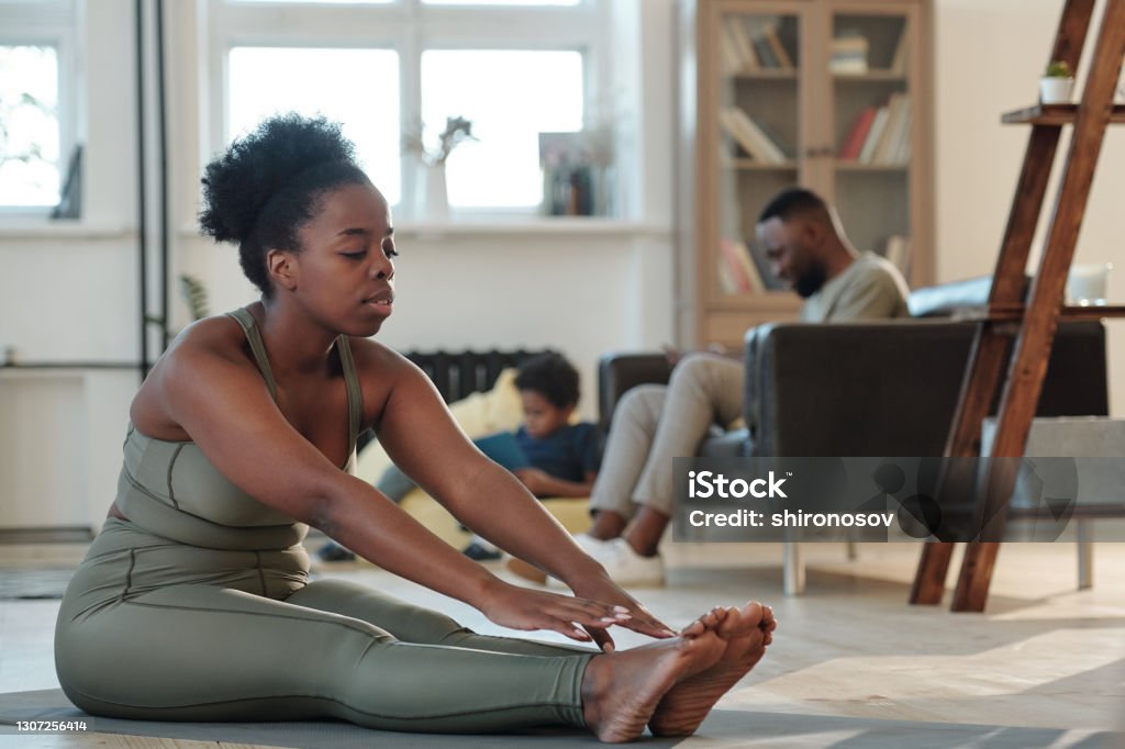 Fit young African woman in tracksuit sitting on the floor of living-room during physical exercise Fit young African woman in tracksuit sitting on mat on the floor of living-room during physical exercise against her husband and little son Exercising Stock Photo