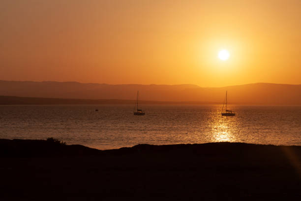 Landscape photograph at sunset, of the Paracas bay, with the silhouette of two sailboats, in the distance, outlined against the orange light, in the Paracas National Reserve, Pisco, Ica, Peru.