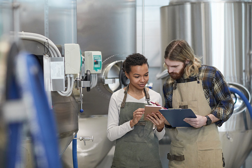 Waist up portrait of two young workers using digital tablet while inspecting production at modern craft brewery, copy space