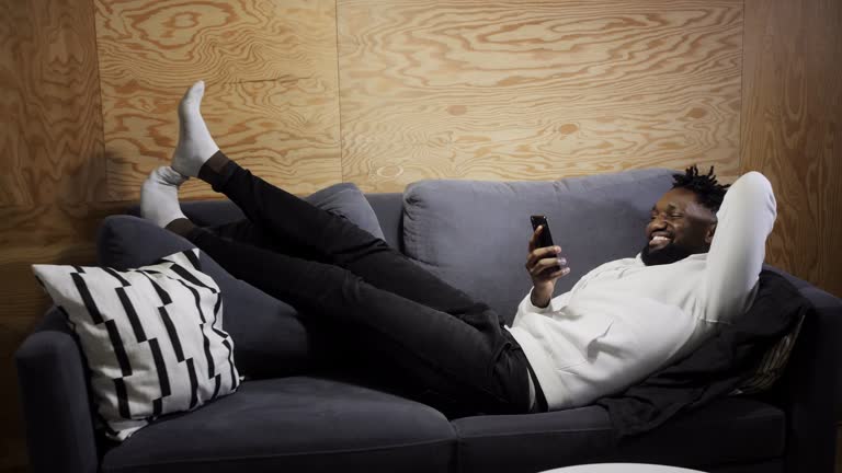 Full length smiling young african ethnicity man sitting on comfy couch, playing online mobile game. Happy mixed race guy looking at smartphone screen, reading pleasant message news, relaxing at home