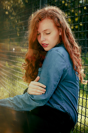 Lovely redhead young model sitting near fence  in rays of sun