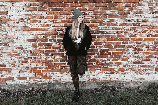 A teenager with long blonde hair standing outside in the background of a brick wall.