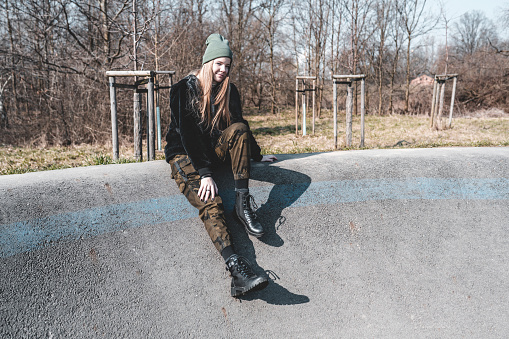 Teenager blond girl wearing camouflage pants and black jacket, sitting on ramp at skatepark on a sunny day.