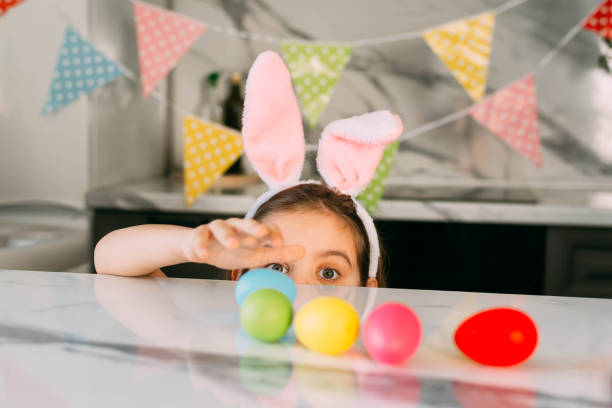 istock Funny girl Kid with Rabbit Ears plays with colored Easter Eggs sitting at the Kitchen table. Holidays Concept. 1307285514