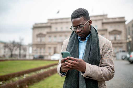 Young African American businessman casually walking around the city, using a smart phone to remotely take care of his business, having a worried look on his face