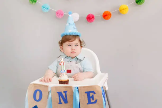 Cute adorable Caucasian baby boy in blue hat celebrating his first birthday at home. Child kid toddler sitting in high chair looking at cupcake dessert with lit candle. Happy birthday concept.