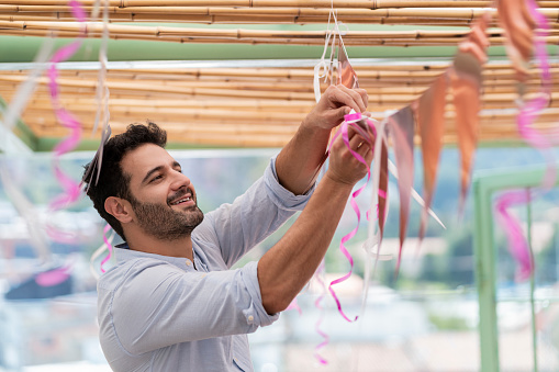 Young man of Latino race, smiles and looks at the camera while assembling the decoration of a birthday party, smiling happily, enjoying the moment