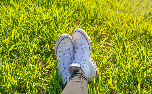 girl's feet in white sneakers sitting in a green meadow in spring on a very sunny day. concept freedom