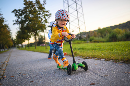 Two caucasian brothers with sports helmets driving balance bike and push scooter outdoors on a sunny day. They are supporting each other.