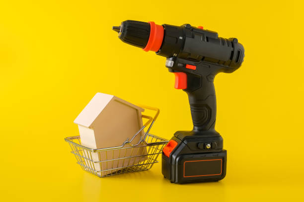 Modern black cordless screwdriver and wooden house in shopping basket Modern black cordless screwdriver, drill with rechargeable battery and wooden house in shopping basket on yellow background, copy space for text, construction or renovation concept chuck wagon stock pictures, royalty-free photos & images