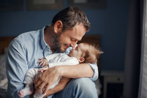 中年の白人の父親は眠っている新生児の女の子にキスをします。揺れる子供の娘の息子を手に持つ親。本物のライフスタイルの子育て父性の瞬間。シングルパパの家族の家庭生活。 - 35 39歳 写真 ストックフォトと画像