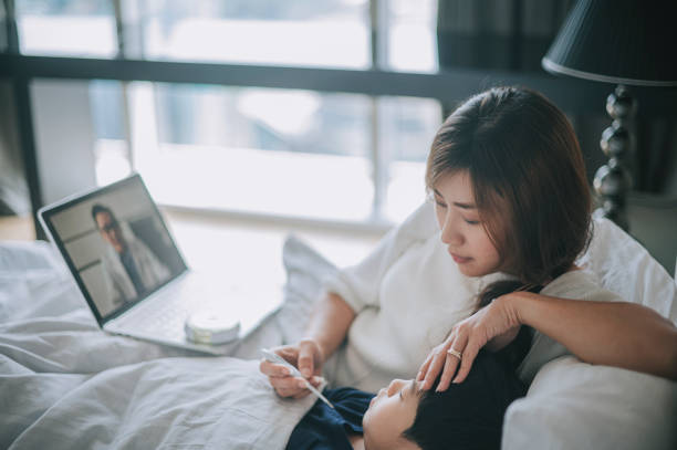 mère chinoise asiatique de télémédecine communiquant avec son docteur utilisant l’ordinateur portatif sur le bâti à côté de son fils dans la maladie - domestic life sadness family bed photos et images de collection