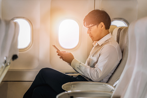 asian businessman in white shirt sitting by window in airplane using smartphone during business trip traveling